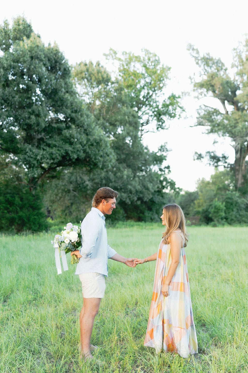 A couple holds hands in a sunlit meadow as they face each other, surrounded by tall grass and trees, capturing a quiet and emotional pregnancy reveal moment.