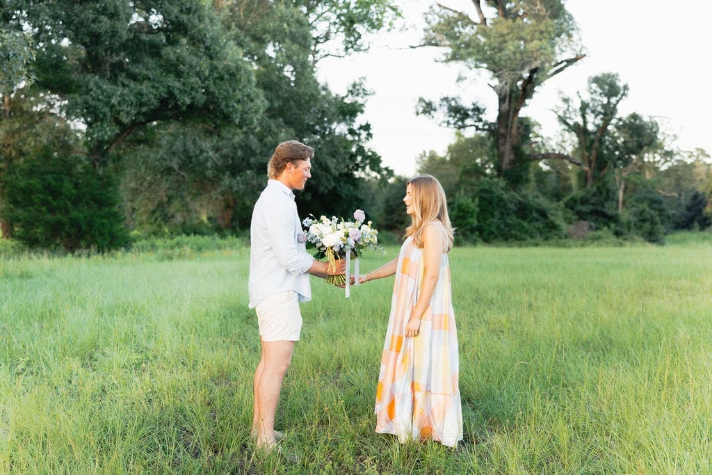 A couple smiles at each other while standing several feet apart in a grassy field, the moment feeling calm, candid, and full of anticipation.