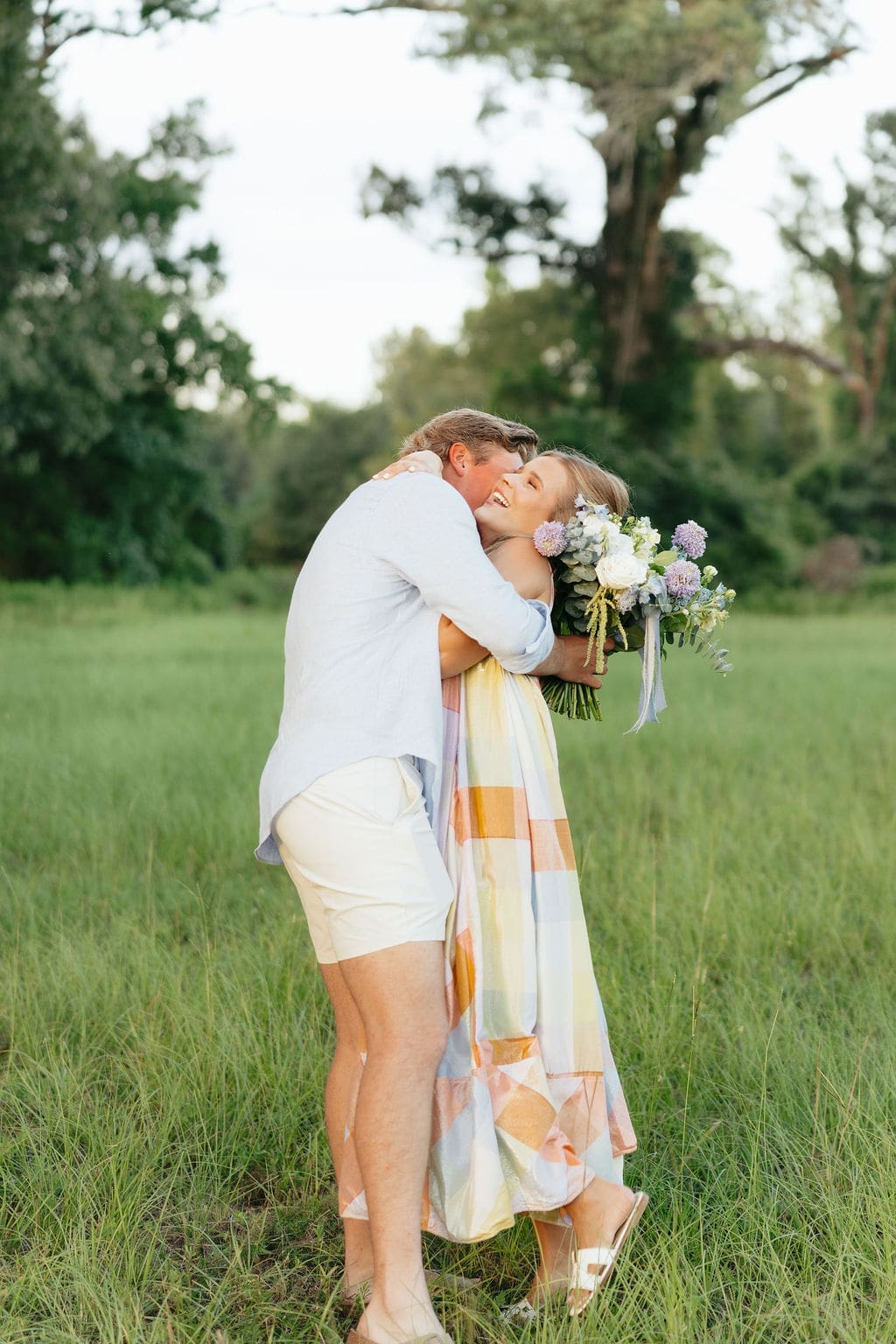 A couple embraces while holding a bouquet in an open field, sharing a joyful reaction that reflects relaxed, documentary-style gender reveal photo ideas outdoors.