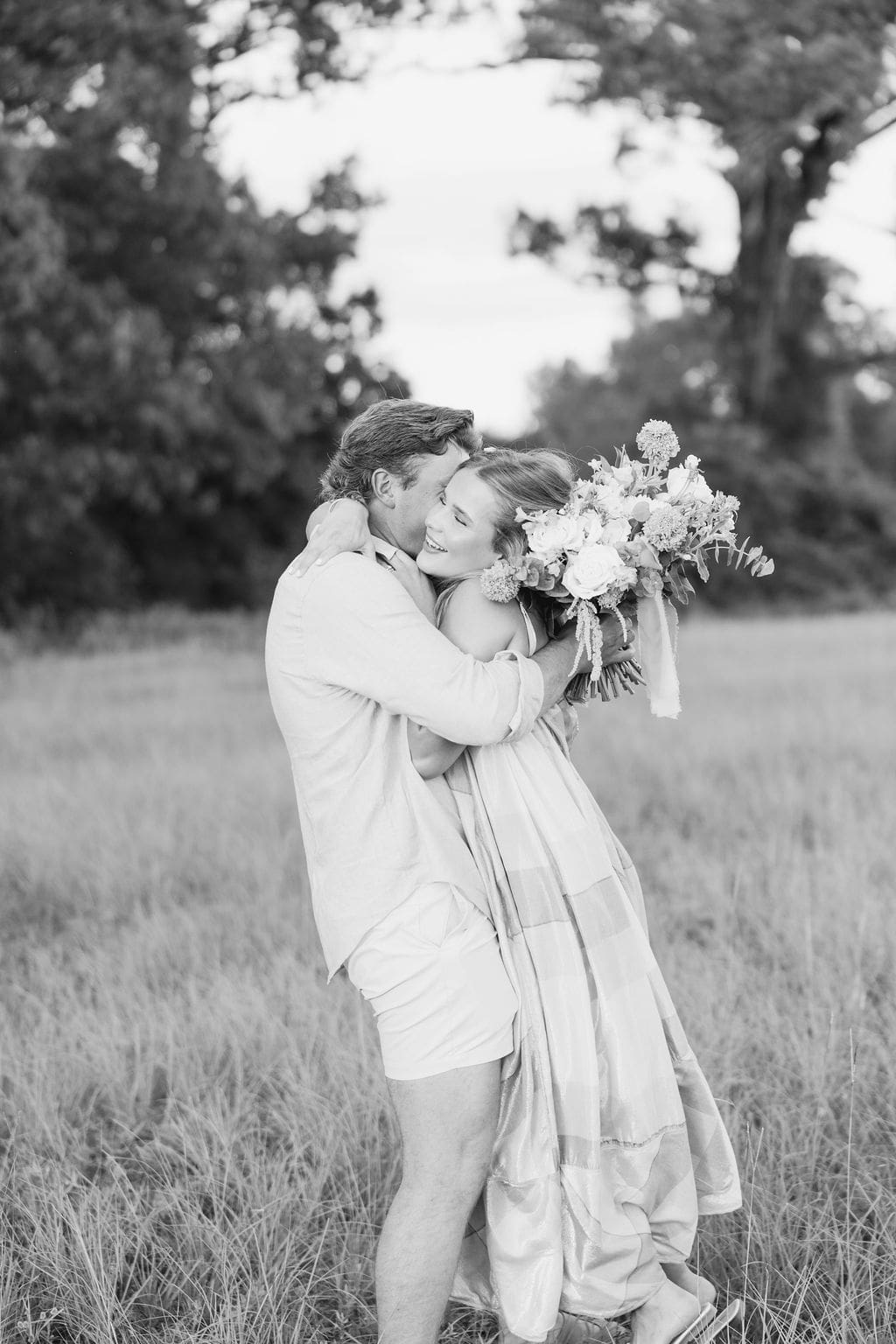 A couple embraces while holding a bouquet in an open field, sharing a joyful reaction that reflects relaxed, documentary-style gender reveal photo ideas outdoors.