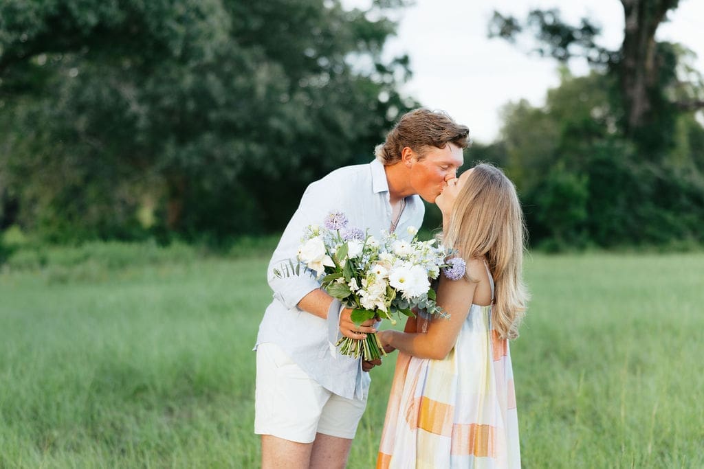 A wide shot of a couple kissing in a field with their gender reveal bouquet in the husbands hands.