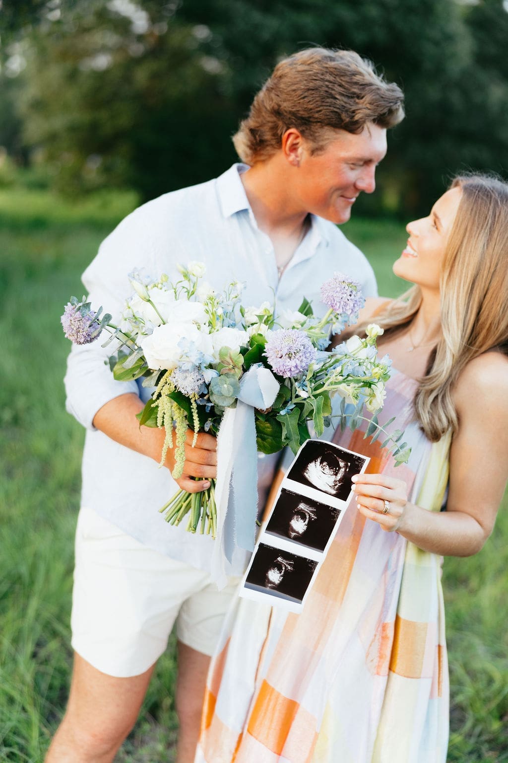 A close-up of the couple smiling at each other while holding a bouquet and ultrasound photos, framed by soft greenery and natural light.