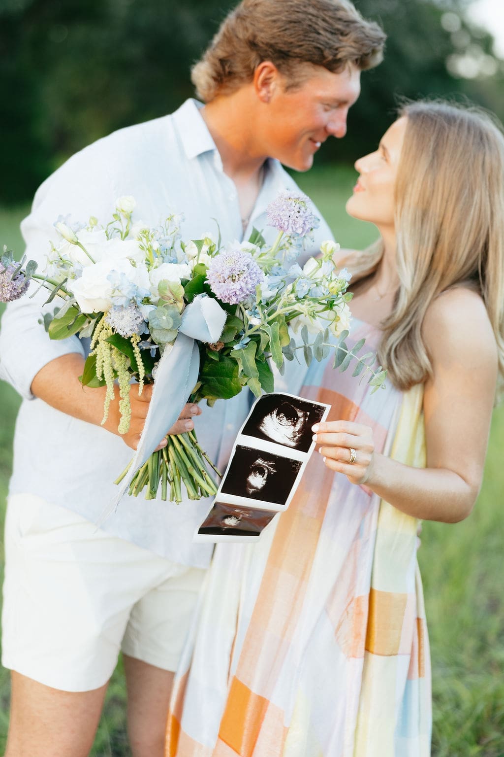 A close-up of the couple smiling at each other while holding a bouquet and ultrasound photos, framed by soft greenery and natural light.