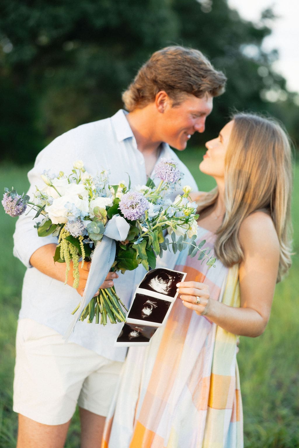 A close-up of the couple smiling at each other while holding a bouquet and ultrasound photos, framed by soft greenery and natural light.