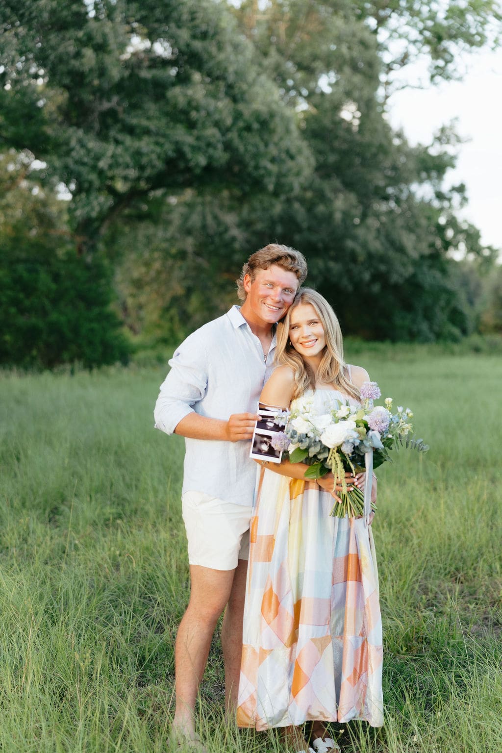 Gender reveal photo ideas: Husband and wife hugging in an open field with their sonogram + floral bouquet with the baby's gender.
