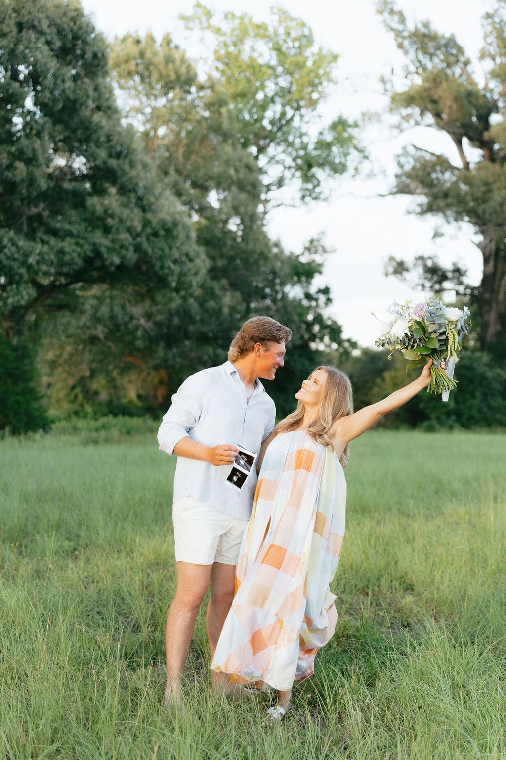 A couple walks through a grassy field the wife lifts a bouquet in celebration, showcasing playful gender reveal photo ideas with movement and joy.