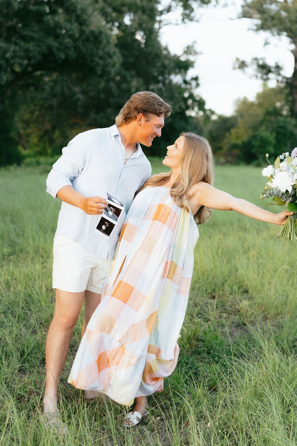 A couple walks through a grassy field the wife lifts a bouquet in celebration, showcasing playful gender reveal photo ideas with movement and joy.