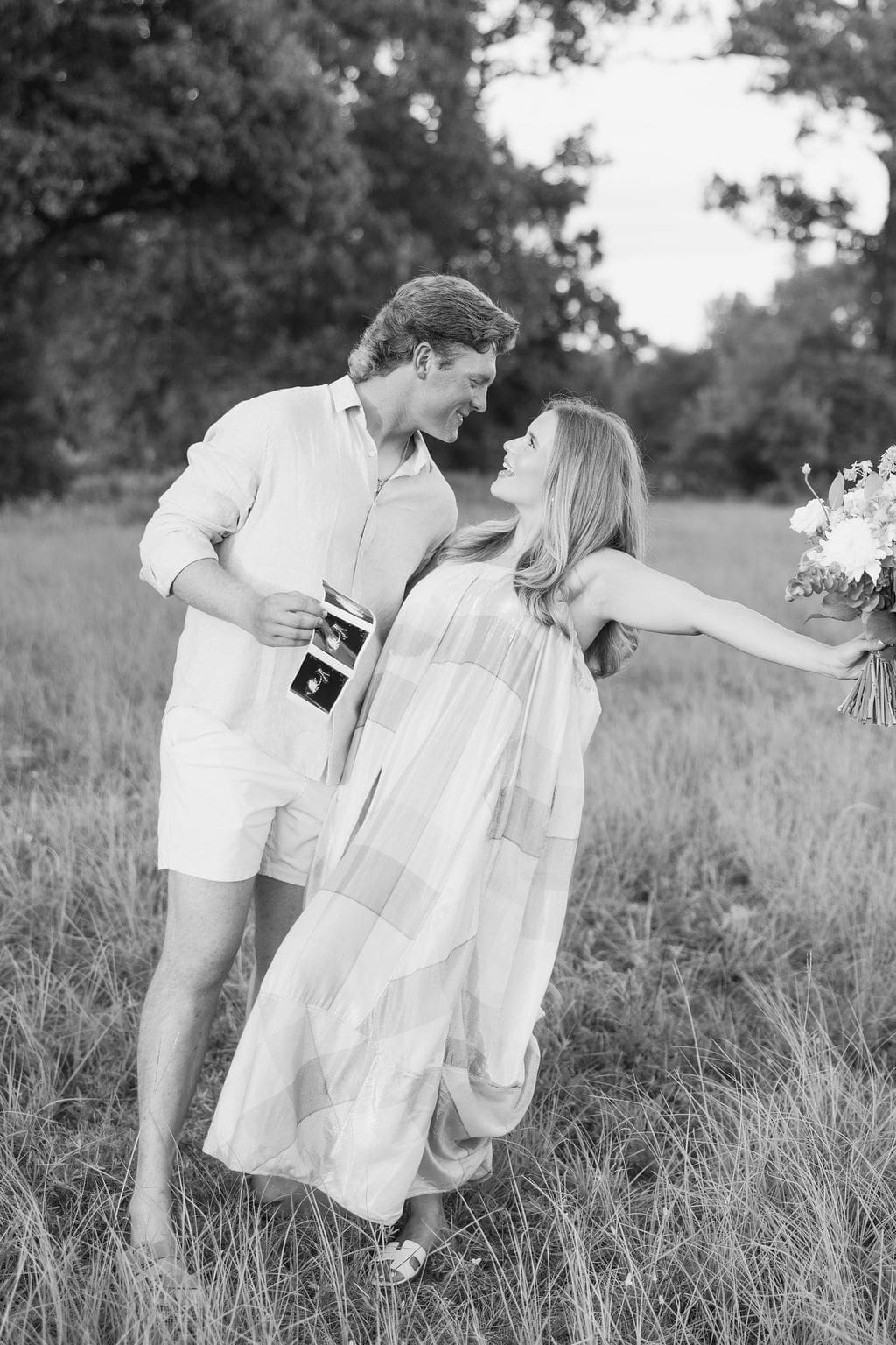 A couple walks through a grassy field the wife lifts a bouquet in celebration, showcasing playful gender reveal photo ideas.