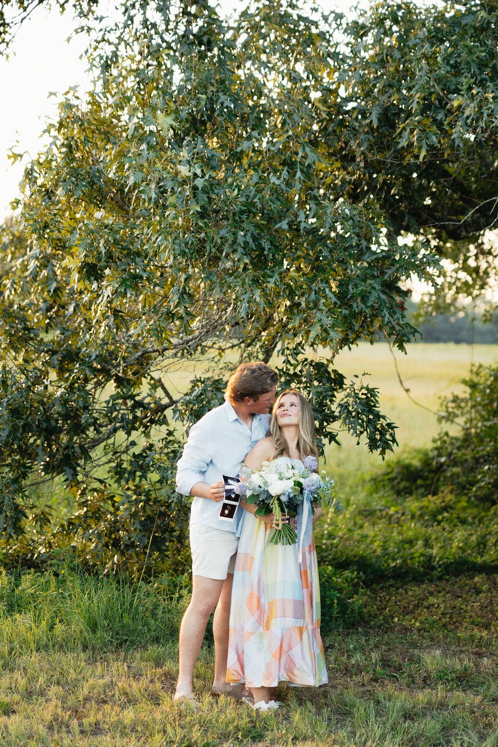 Gender reveal photo ideas: A couple holding a bouquet with the gender reveal color and their sonogram photos at sunset in an open field.