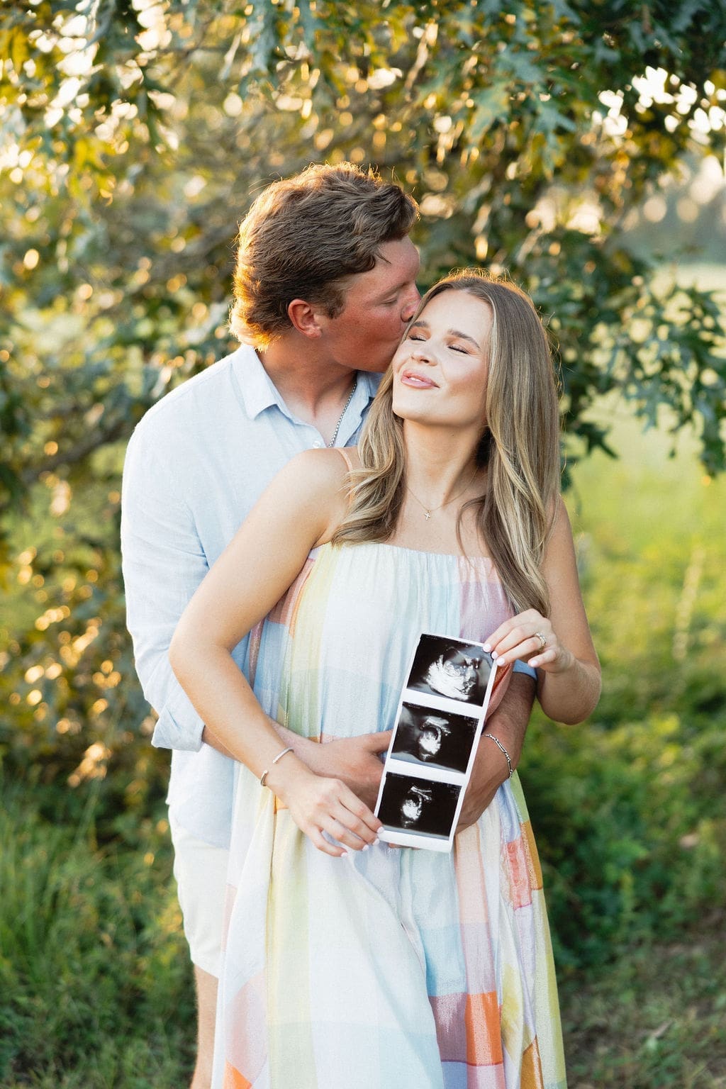A close-up of the husband kissing his wifes forehead while she is holding a bouquet and ultrasound photos, framed by soft greenery and natural light.