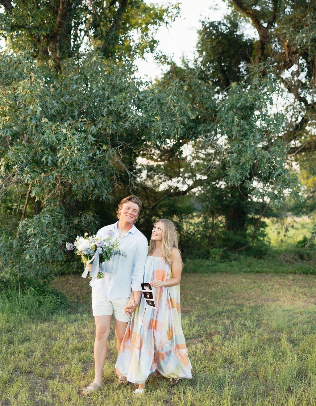 A couple walking in a field with a gender reveal floral bouquet + their sonogram for their gender reveal photos.
