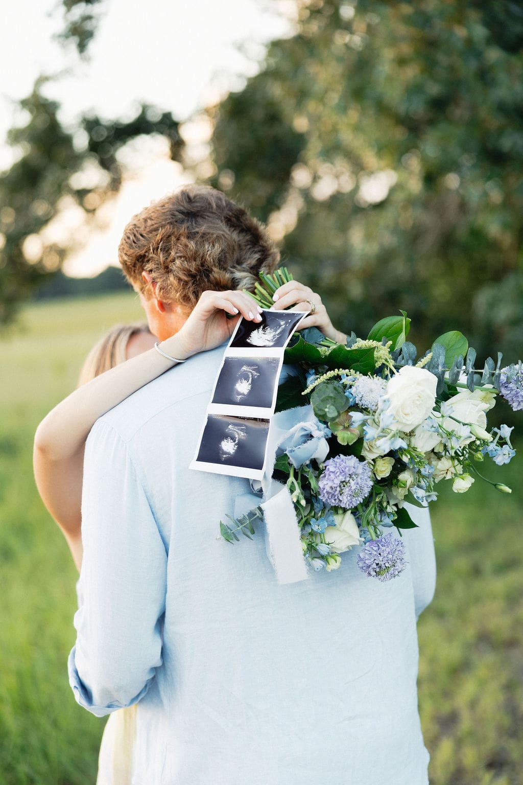 A couple hugs closely in a field as ultrasound photos rest against a bouquet, capturing an intimate and emotional pregnancy announcement moment.