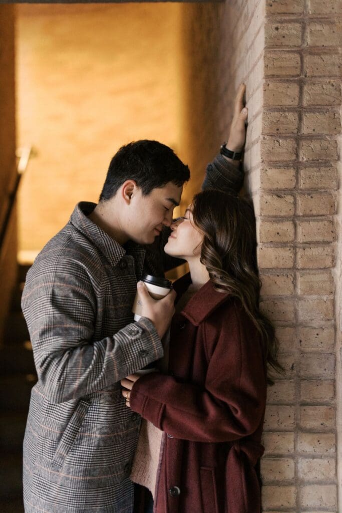Romantic Engagement Photo Ideas: Couple leaning against a wall with their noses touching while holding coffees.