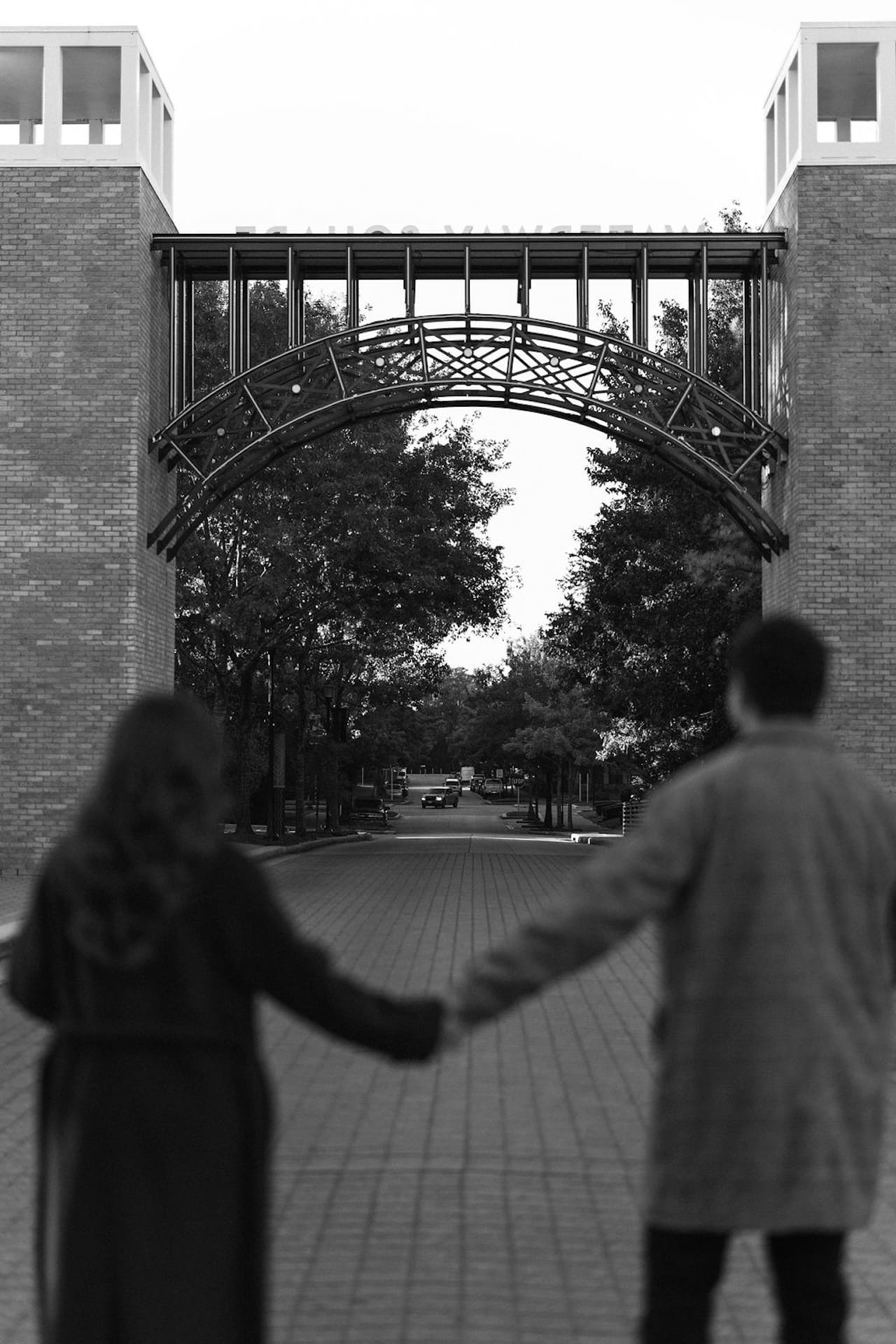 A black and white photo of an engaged couple standing hand in hand in the street. Them blurred in the foreground and the trees and a bride in focus in the background.