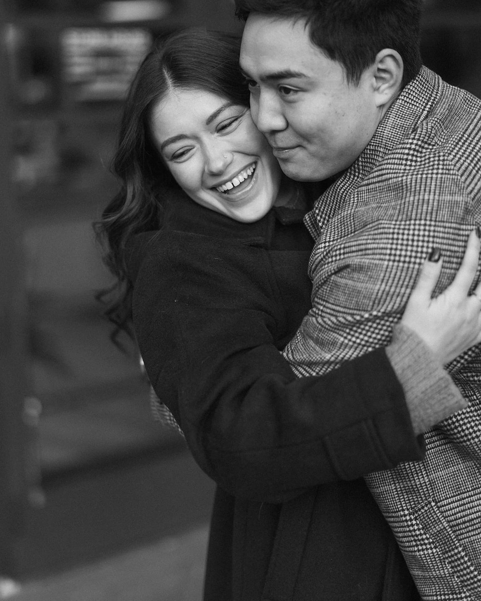 Black and white phtoo of the engaged couple hugging into each other and smiling off into the distance.