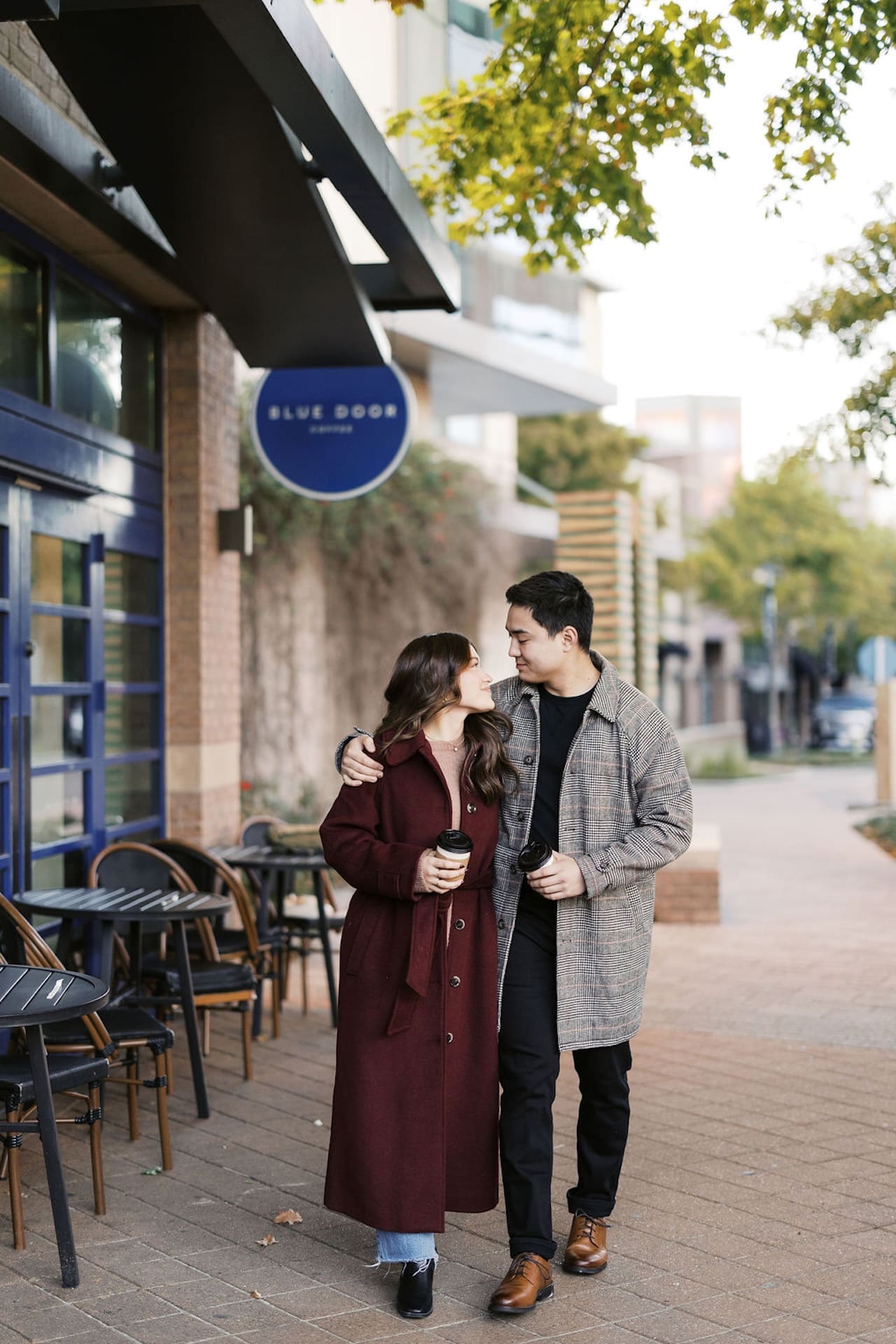 Engaged couple walking along a sidewalk holding coffees during their morning engagement shoot