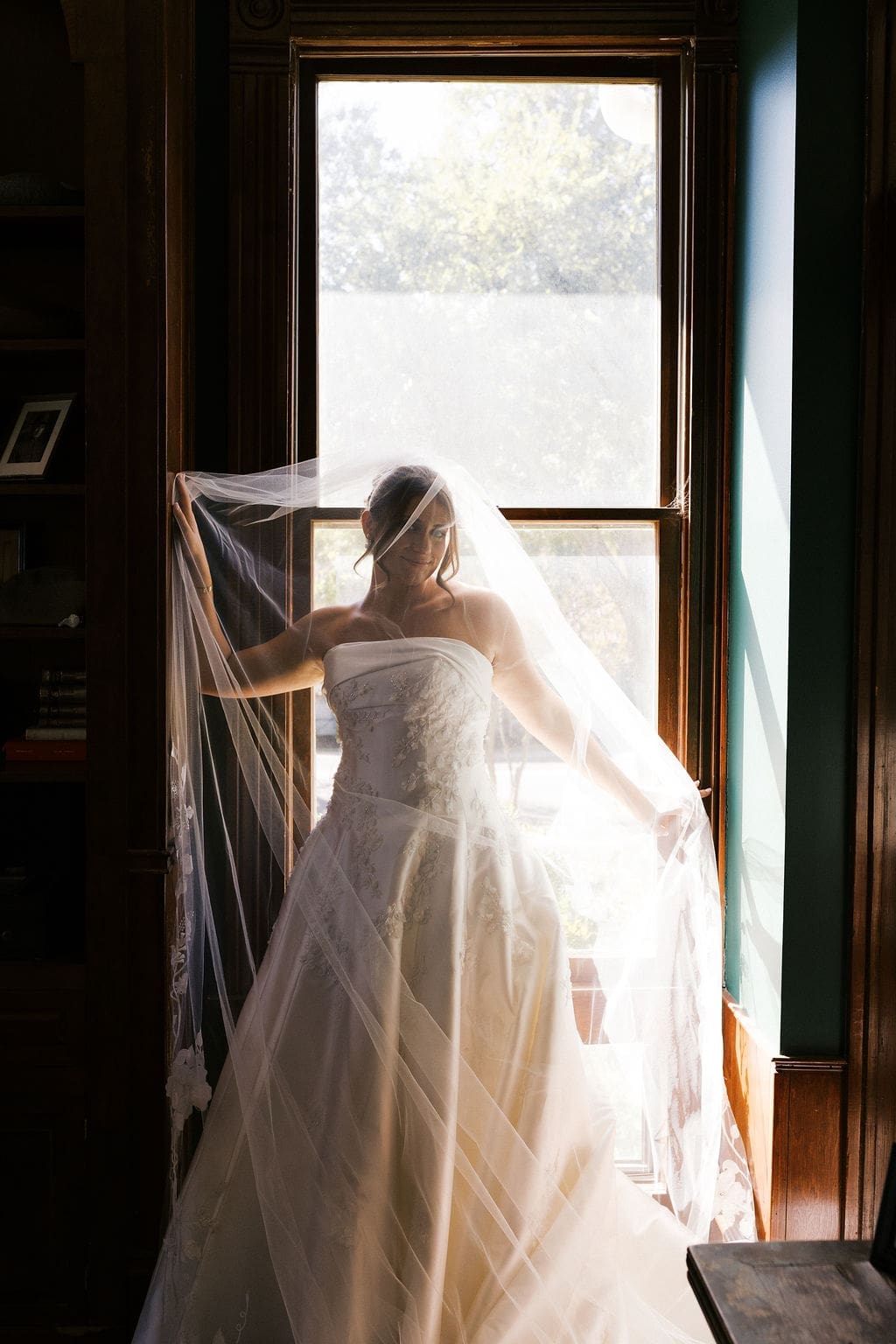 An editorial bride portrait of the bride with her veil draped around her as she leans against a window was the sun shines behind her and through her veil.