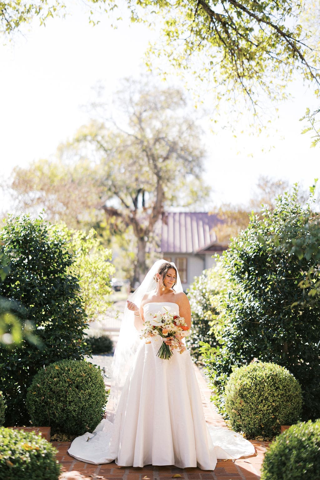 The bride standing in the gardens at Woodbine Mansion smiling softly and holding her wedding bouquet and playing with her veil.