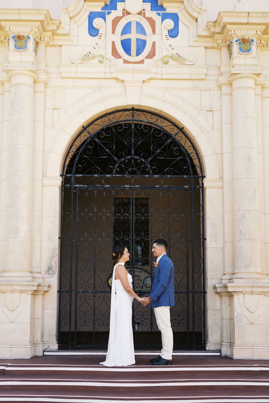 Couple facing each other and holding hands in front of tall iron gates and decorative courthouse architecture during their intimate elopement ceremony.