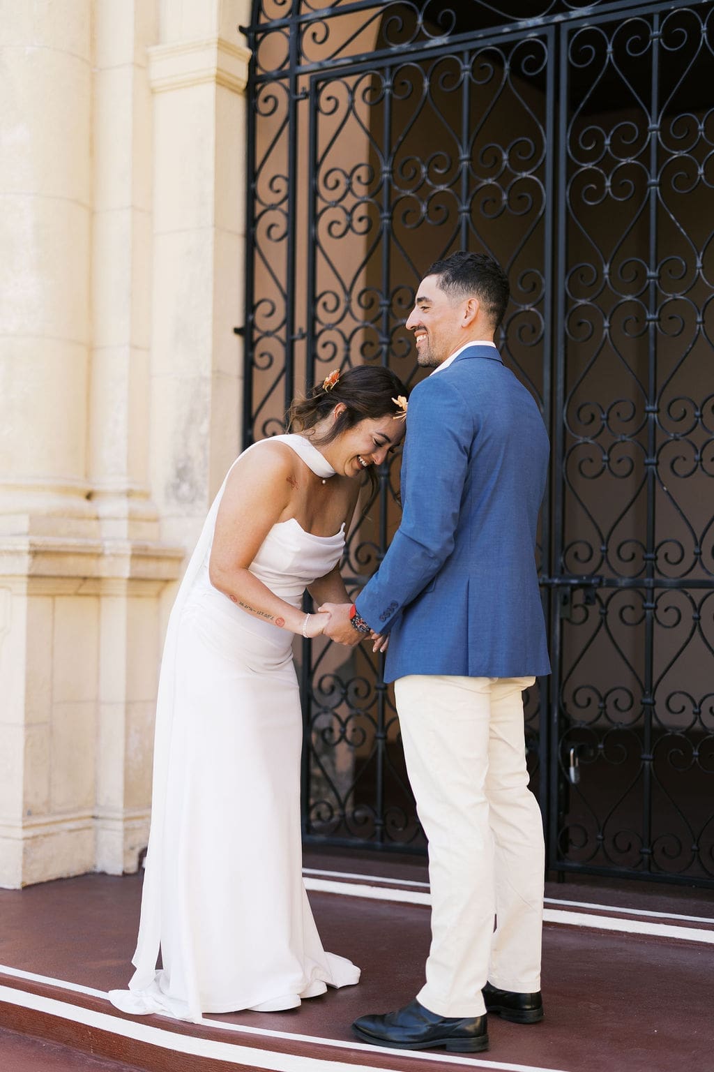 Bride and groom laughing together while holding hands in front of ornate courthouse doors, sharing a candid moment during their elopement portraits.