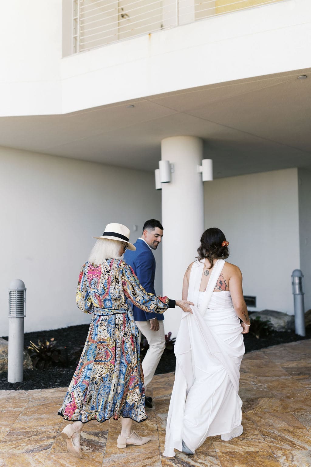 Bride, groom, and a family member walking together outside a modern building, with the bride’s dress train being held and the couple heading to their destination elopement courthouse ceremony.