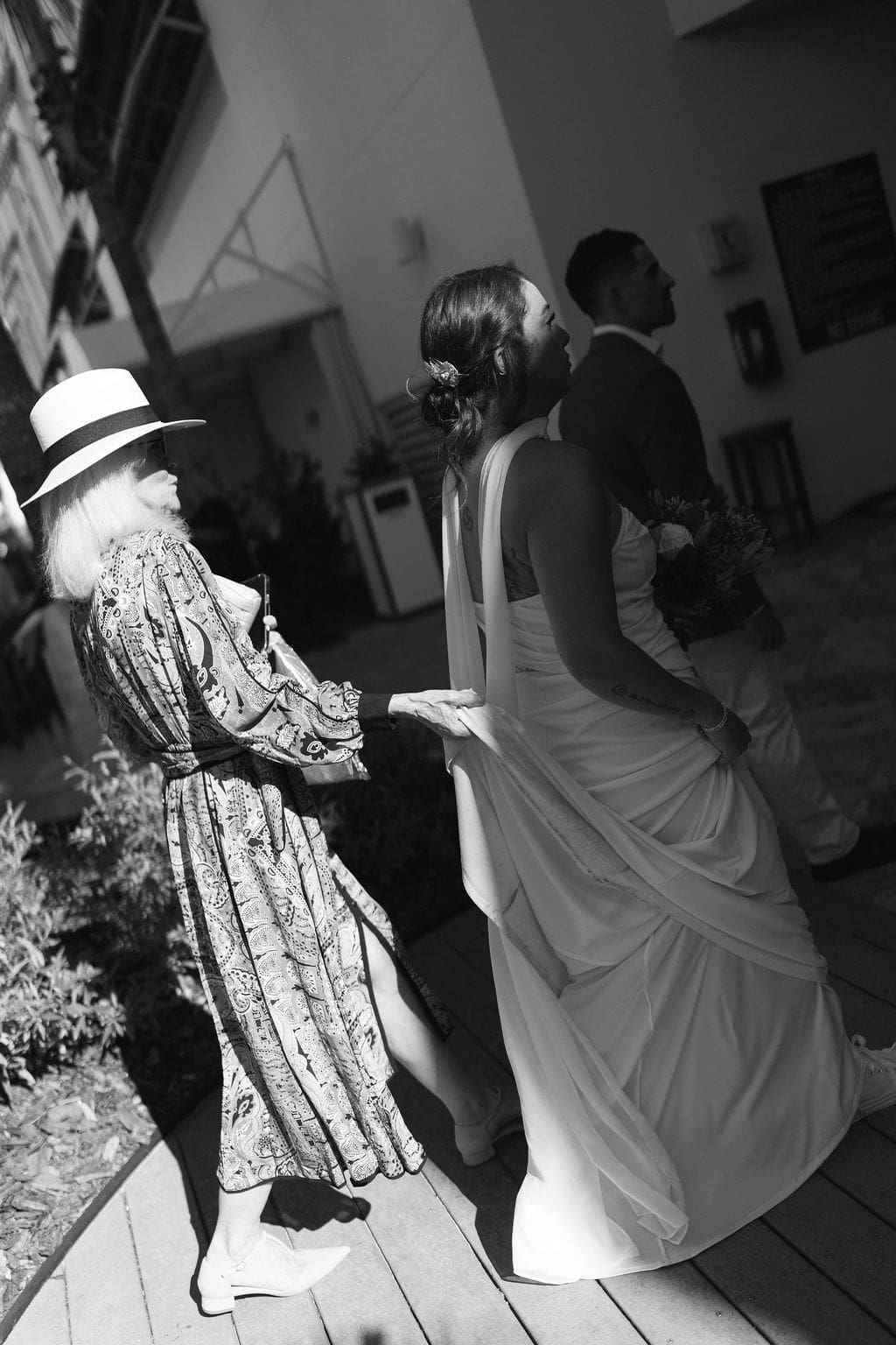 Black and white photo of a woman adjusting the bride’s dress train as the couple walks toward their ceremony, capturing candid elopement moments with family.