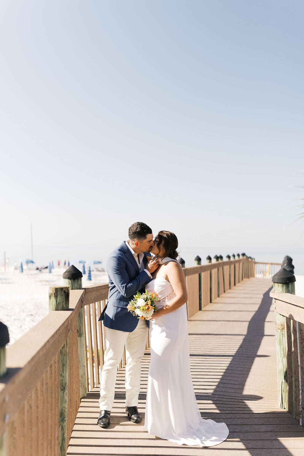 Bride and groom kissing on a beach boardwalk while holding a bouquet, with bright sun and ocean scenery behind them.