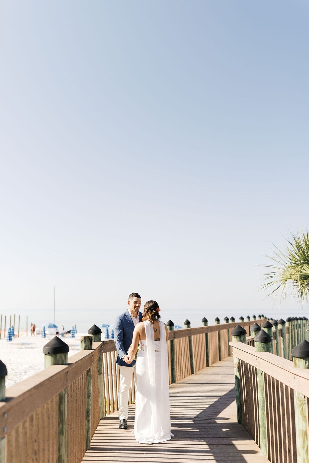 Couple holding hands on a wooden boardwalk with the ocean and beach umbrellas in the background, sharing relaxed destination elopement portraits.