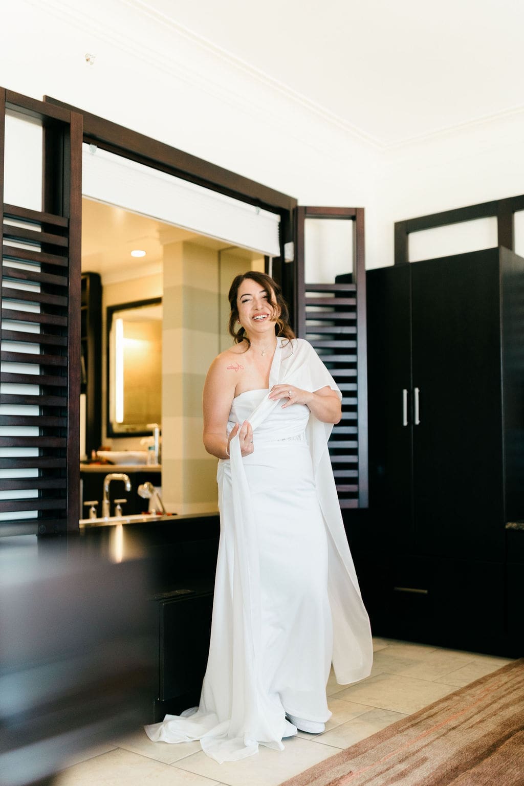 Bride laughing while standing in a modern hotel suite wearing her wedding dress before her destination elopement.