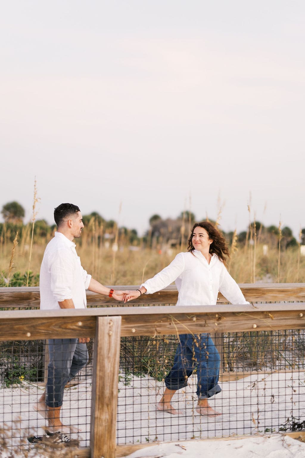 Couple holding hands and walking along a wooden boardwalk through coastal dunes, wearing casual white outfits for their Florida beach elopement portraits.