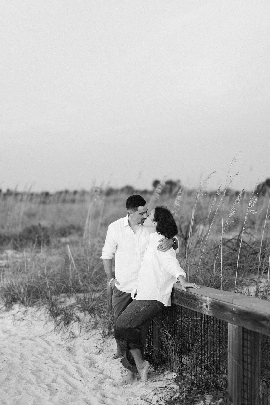 Couple leaning against a wooden railing in coastal dunes, sharing a quiet embrace during their destination elopement beach session.