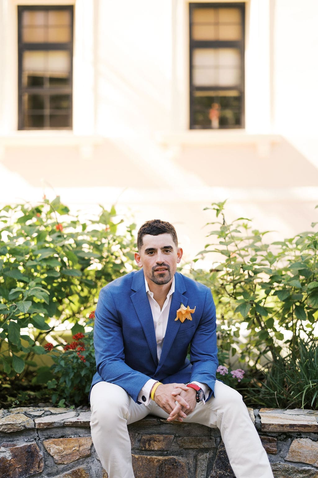 Groom sitting on a stone ledge in a blue suit with a boutonniere, posing in a garden courtyard during destination elopement portraits.
