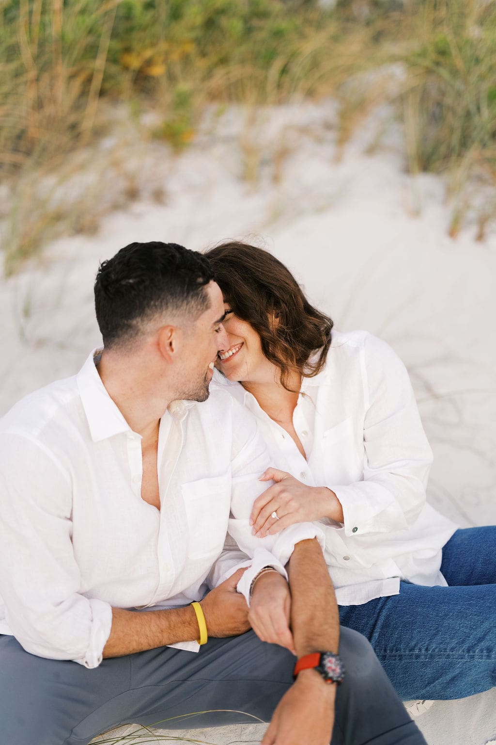 Couple laughing together while sitting in the sand near sea oats, capturing playful moments from their destination elopement portrait session.