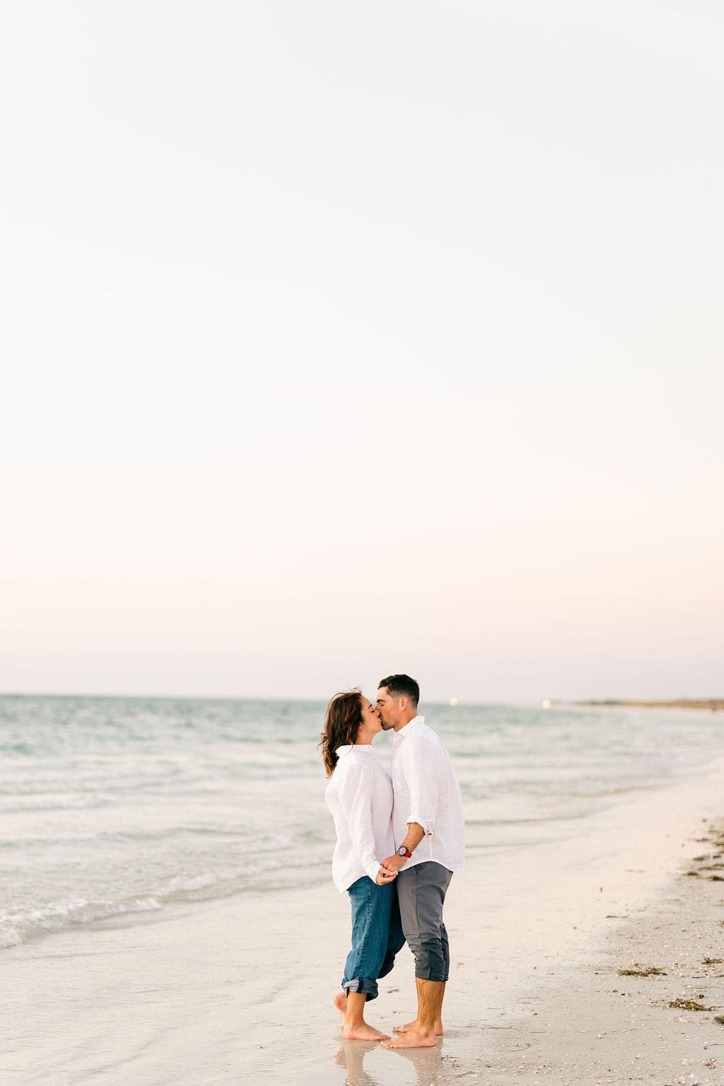 Bride and groom holding hands and kissing by the shoreline with calm ocean waves behind them during sunset beach portraits.