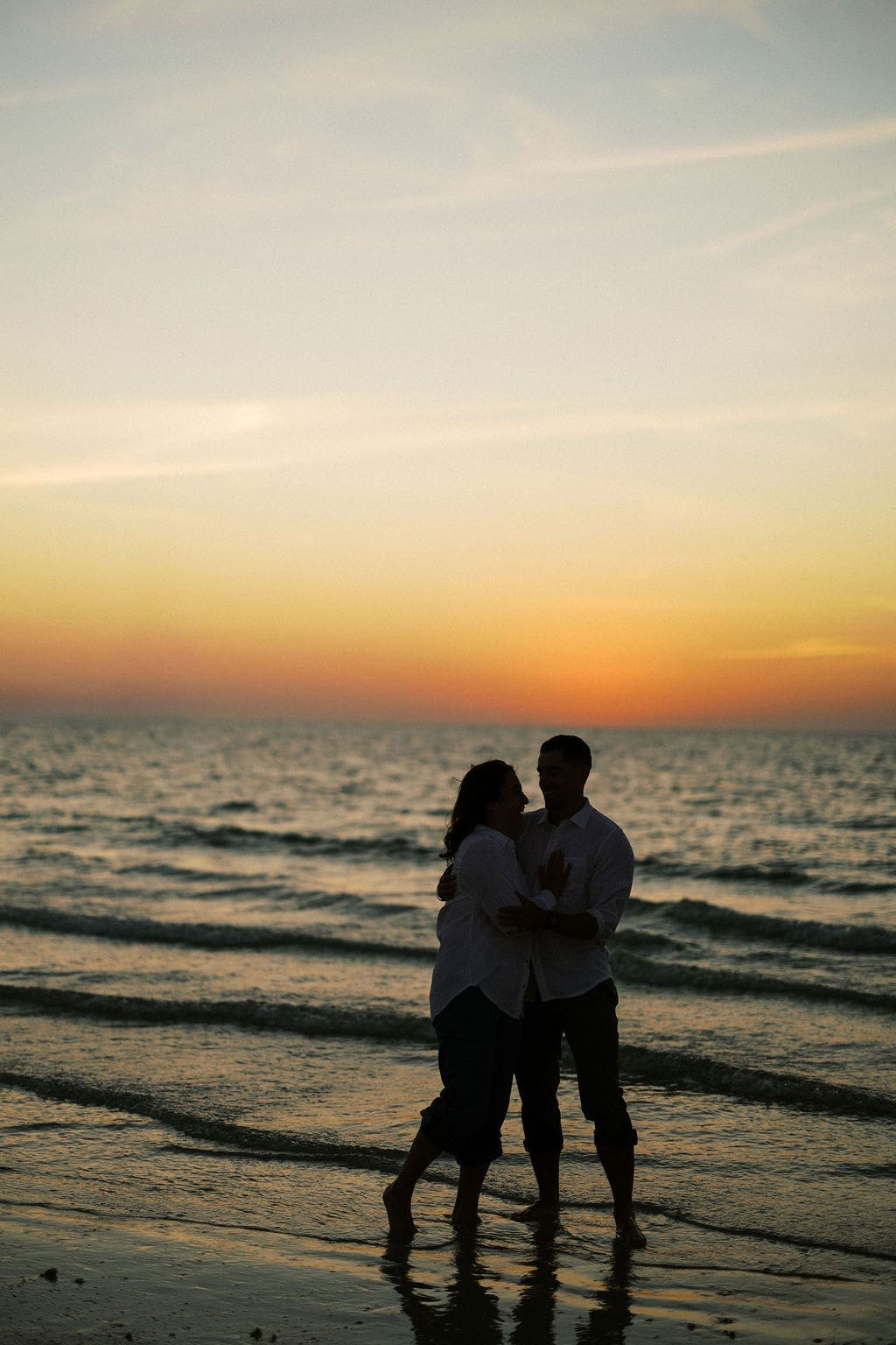 Couple embracing and laughing on the beach at sunset with waves and colorful sky in the background.
