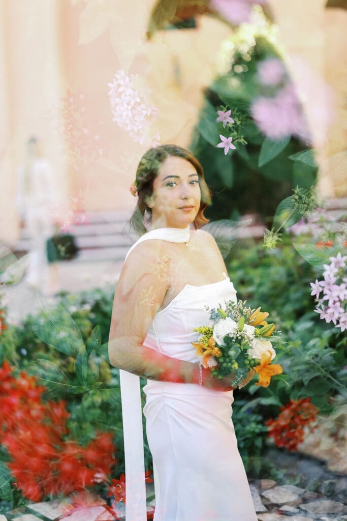 Bride holding a bouquet in a lush garden, photographed through soft floral foreground for a dreamy, layered portrait.