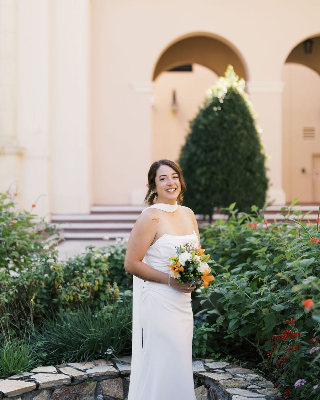Bride smiling and holding a colorful bouquet in a white wedding dress in front of a historic courthouse building with arches and greenery.