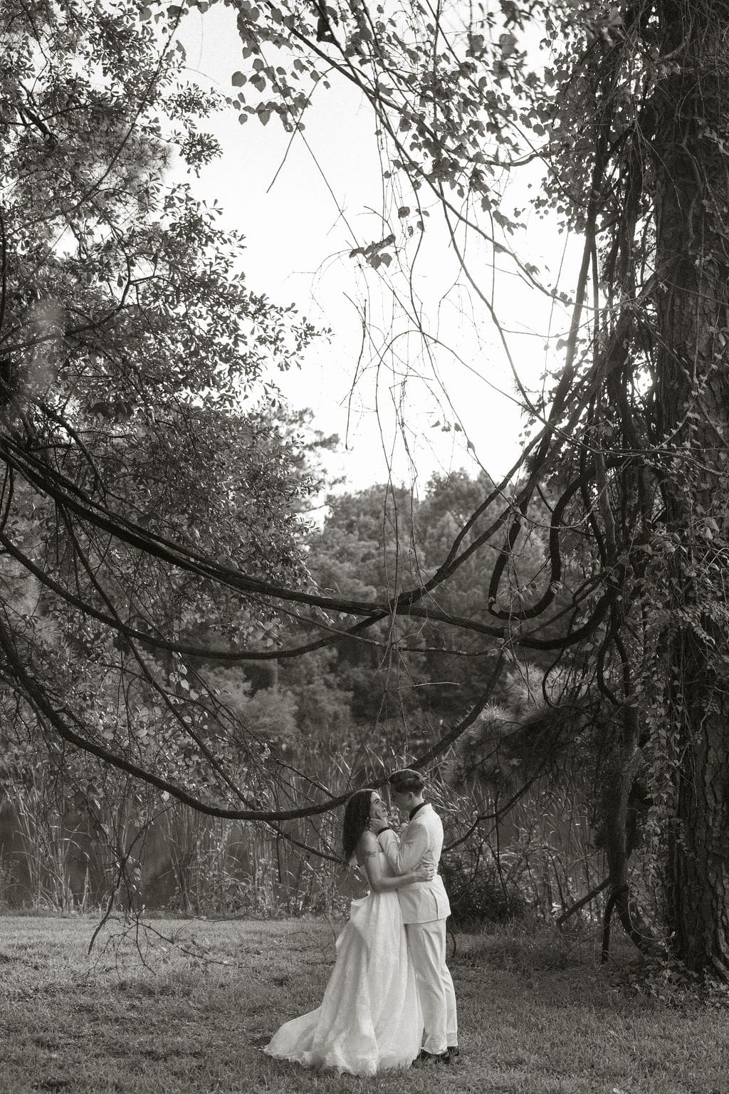 Timeless documentary wedding photos of the bride and groom standing on the lawn with trees all around them as the groom leans in to kiss the bride.