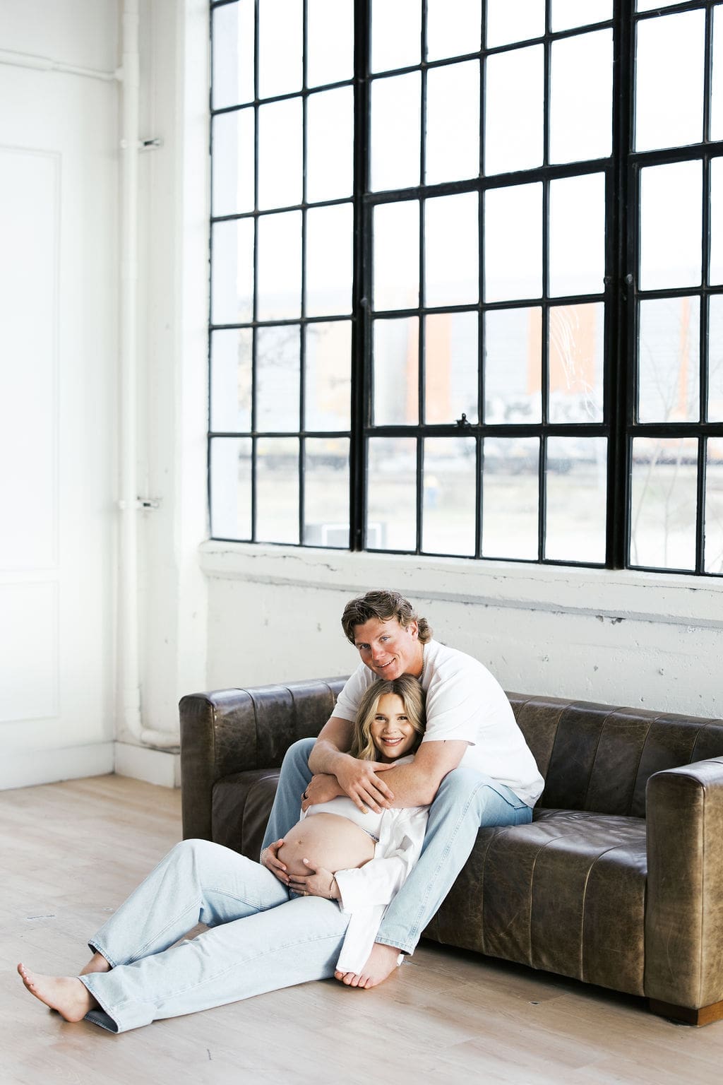 A studio maternity photo of the husband sitting on a couch while the wife sits between his legs while he hugs around her shoulders and she holds her belly.
