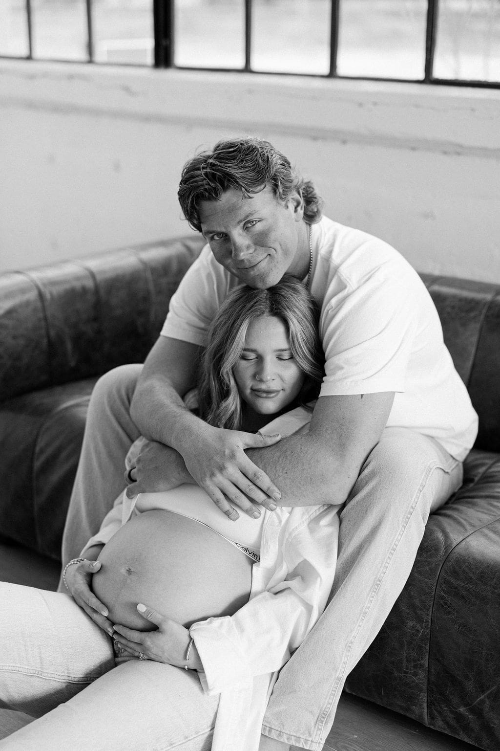 A studio maternity photo of the husband sitting on a couch while the wife sits between his legs while he hugs around her shoulders and she holds her belly.
