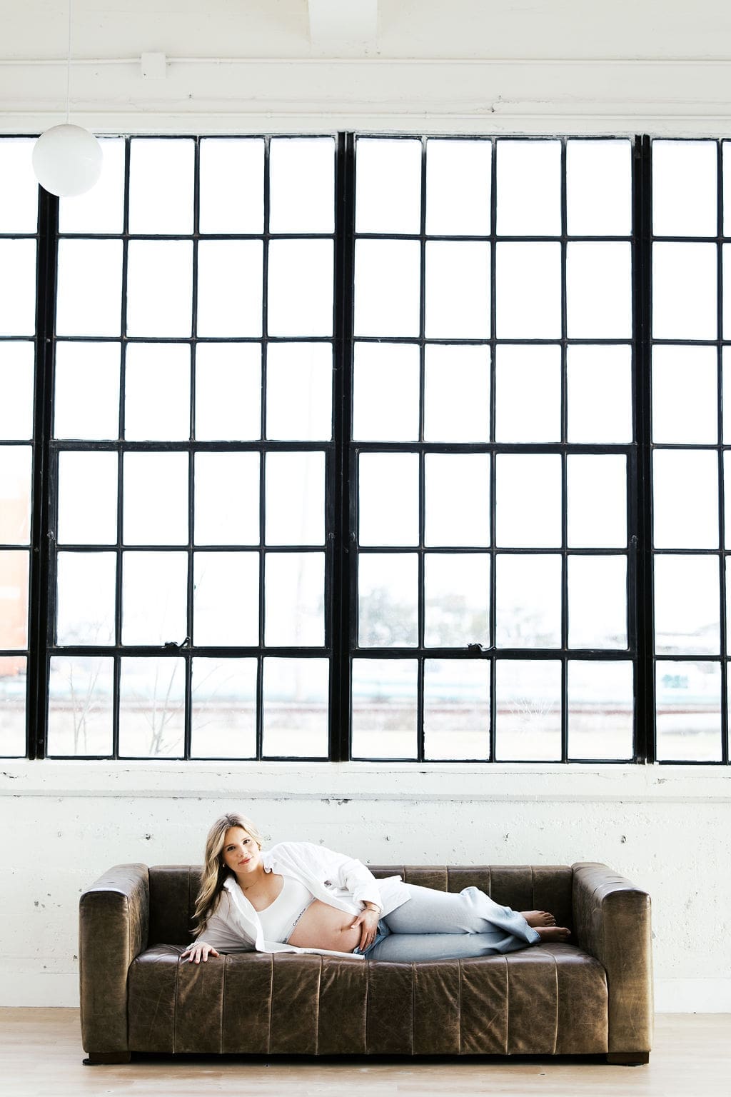 A studio maternity photo of the mom laying down on the brown leather couch with windows behind her as she holds her belly
