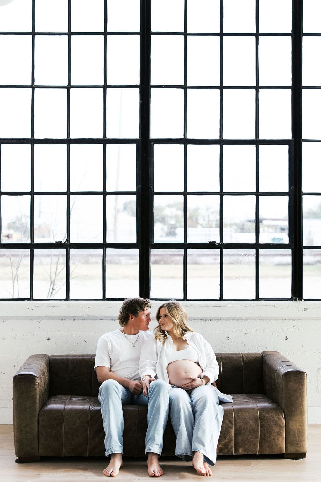 A couple sitting on a leather couch in a studio while smiling at each other for a candid studio maternity photo.