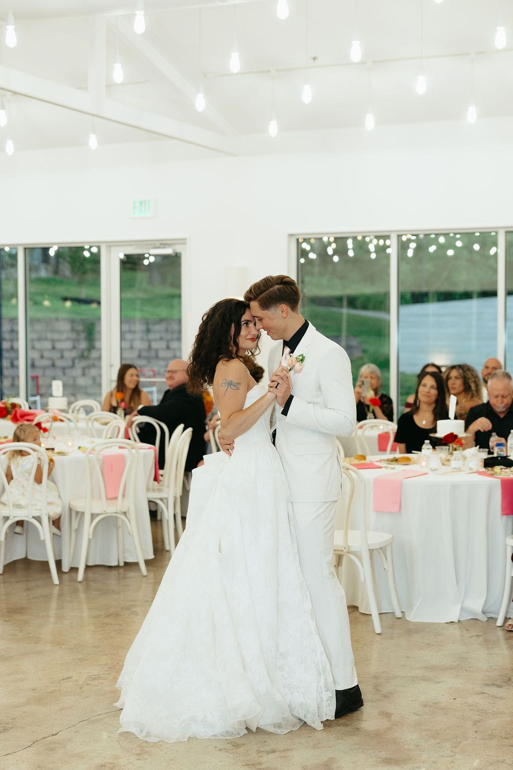 Bride and groom share their first dance in a bright white reception space with string lights overhead, surrounded by guests watching and smiling, captured in romantic documentary wedding photos.