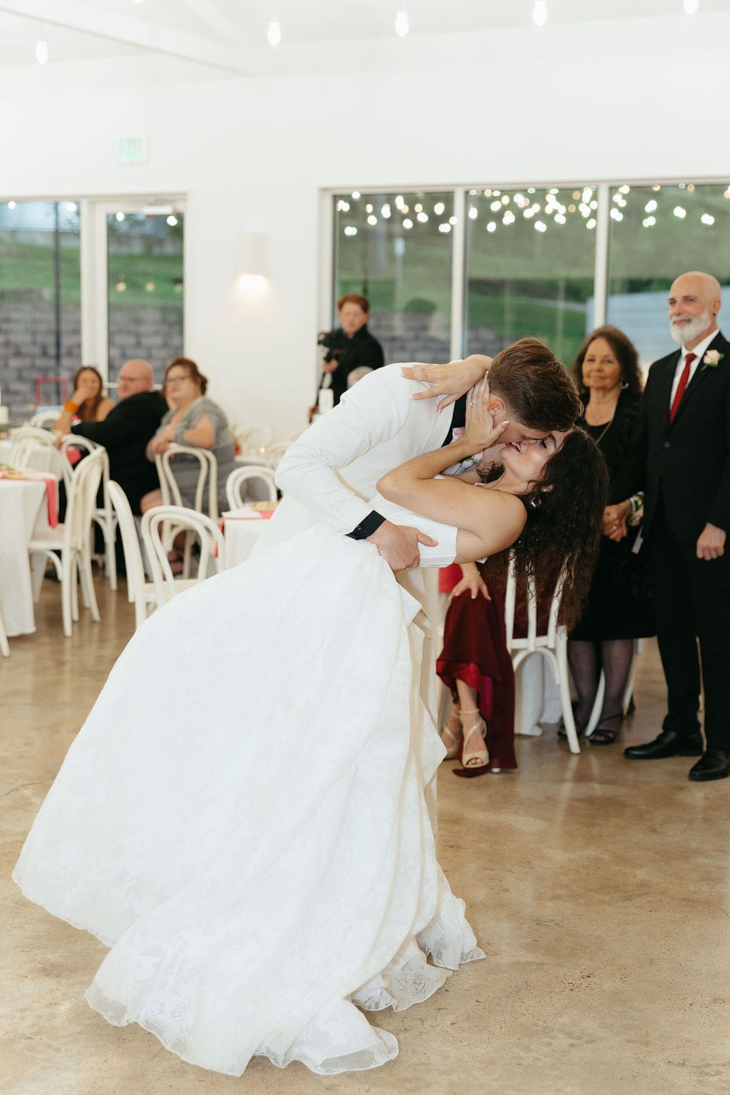 Bride dips backward as the groom kisses her during their first dance while family looks on in the background at the indoor reception.