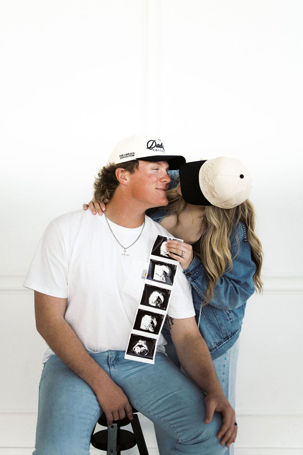The husband sitting on a stool while the mom holds the sonogram over his shoulder and smiles at her husband during their studio maternity photoshoot.