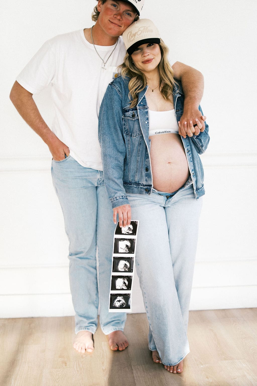 A studio maternity photo of the husband hugging around the wife as she holds his hand on her shoulder while she holds their sonogram.