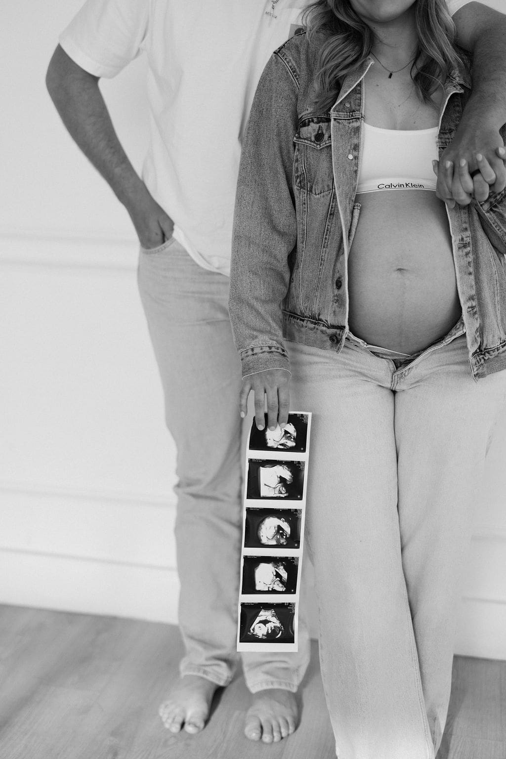 A black and white studio maternity photo of the husband hugging around the wife as she holds his hand on her shoulder while she holds their sonogram.