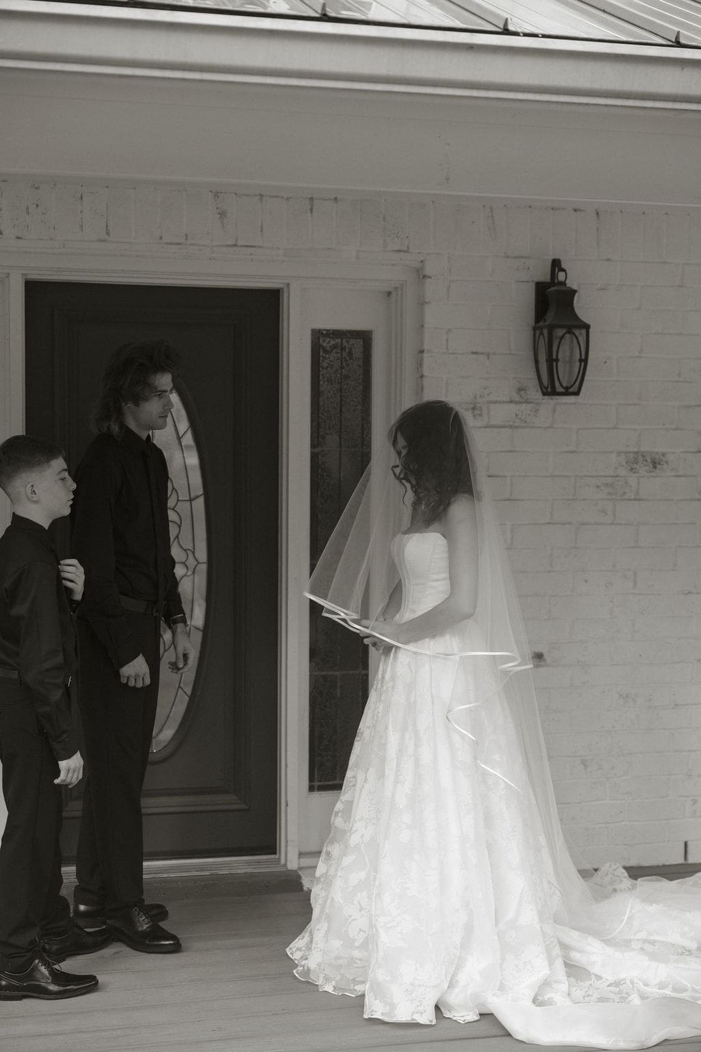 Bride stands under her veil on the porch facing two young men dressed in black, sharing a quiet pre-ceremony moment.