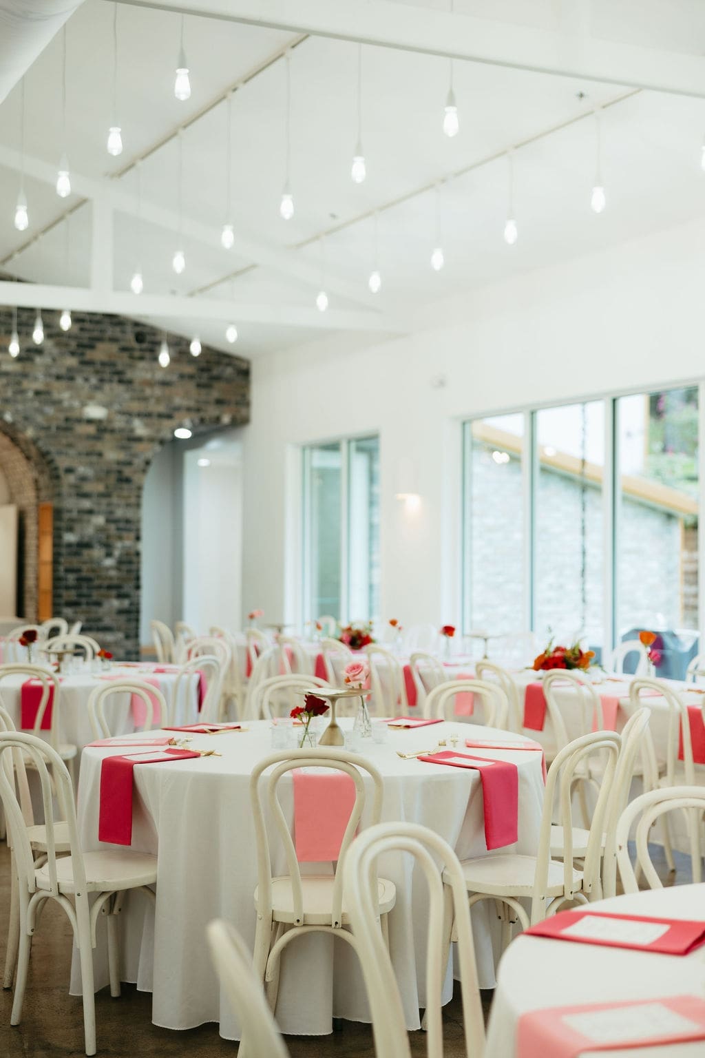 Wide view of the wedding reception space with round tables, pink napkins, and hanging lights, styled simply and naturally for timeless documentary wedding photos.