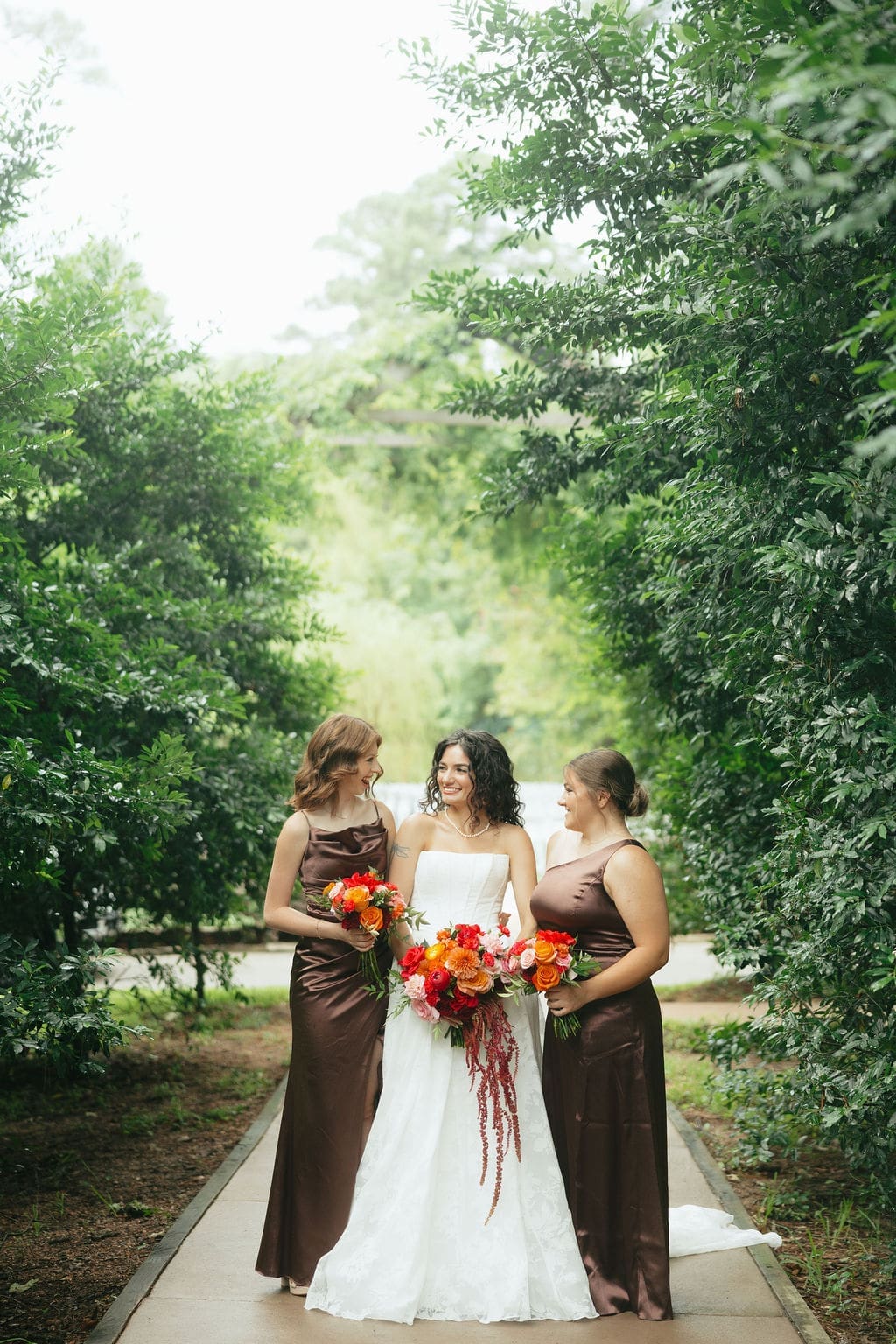 A photo of the bride and her bridesmaids standing along a tree-lined aisle while holding colorful floral bouquets for candid wedding portrait photos. A stunning example of documentary wedding photos.